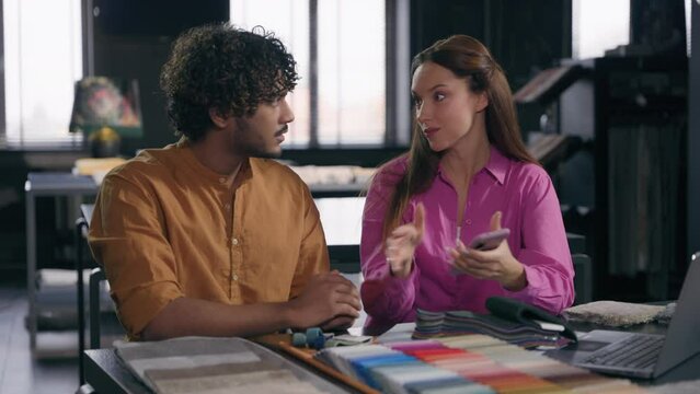 Multiracial couple creative fashion designers choose fabric textile cloth materials palette in atelier Caucasian woman saleswoman and Indian man customer using mobile phone for house renovation design