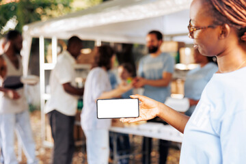 Image showcasing young black woman horizontally holding a digital phone with white screen copyspace display at an outdoor food bank. Volunteers making a difference in hunger relief efforts. - obrazy, fototapety, plakaty