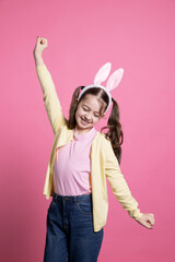 Happy toddler with pigtails and fluffy ears fooling around in the studio, playing and acting silly in front of the camera. Joyful little girl dancing with confidence over pink background.