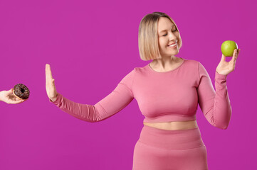 Pretty young woman in sportswear with apple refusing to eat donut on pink background. Weight loss...
