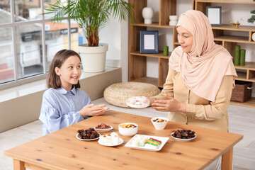 Happy Muslim mother and her daughter sitting at table with traditional sweets in living room. Eid...
