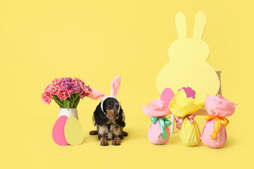 Cute cocker spaniel in bunny ears with Easter gift eggs, paper rabbit and beautiful tulips on yellow background