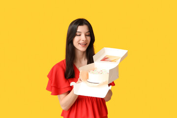 Beautiful young woman holding box with sweet bento cake on yellow background. International Women's Day