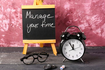 Blackboard with text MANAGE YOUR TIME,  alarm clock and glasses on wooden table against pink...