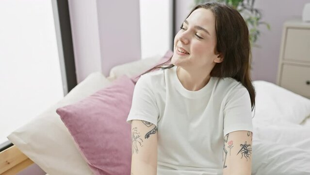 Laughing confidently, young woman in pyjamas radiates happiness, casually sitting on bedroom bed, looking away with natural expression, smiling beautifully.