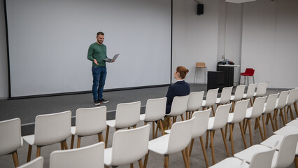 A red-haired Caucasian businesswoman sits in the front row of an empty conference room. Bearded man...