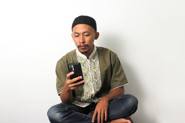 Bored Indonesian Muslim man in koko and peci scrolls through his phone, seeking distraction while...