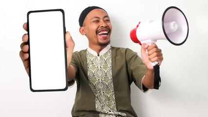Excited Indonesian Muslim man in koko and peci shows a blank phone screen and holds a megaphone,...