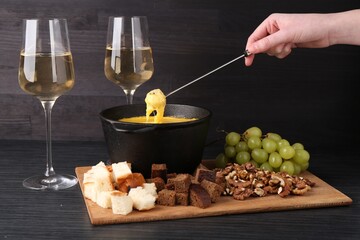 Woman dipping piece of bread into fondue pot with melted cheese at black wooden table, closeup