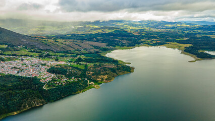 Aerial view of serene Guatavita Lake, lush greenery, quaint town, and mountains under a cloudy sky...