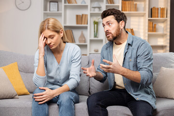Man screaming at his wife sitting on their living room sofa, she demonstrating a headache