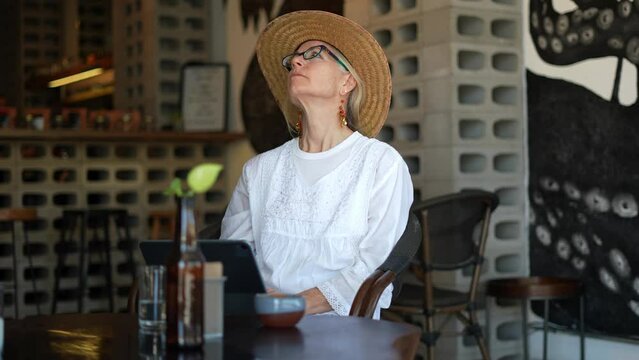 Attractive blond happy mature woman digital nomad using a laptop computer at cafe in in Mexico wearing straw hat contemplating.