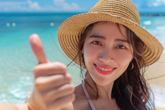 Asian woman with hat at the beach taking a selfie picture doing the thumbs up gesture