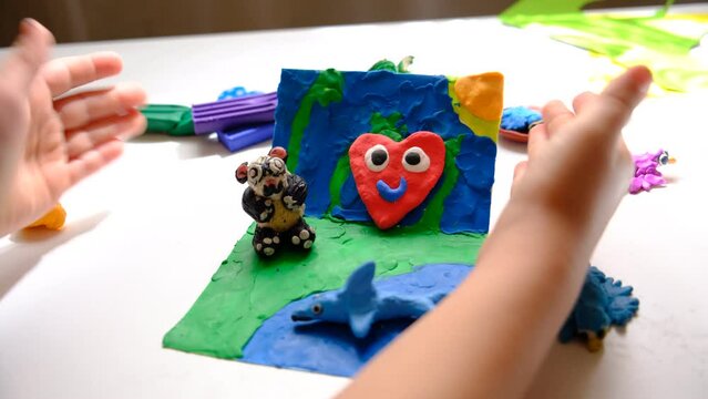 Child smearing colorful plasticine on cardboard and creating fairy tale card with cartoon animals, panda, birds, shark. By spreading, molding and adding texture to plasticine child creating scene