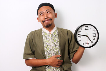 Hungry Indonesian Muslim man in koko and peci holds his stomach and looks longingly at a clock,...