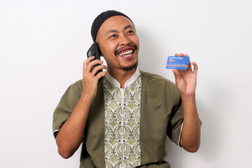 Happy Indonesian Muslim man in koko and peci talks on his phone while holding a credit card, ready...