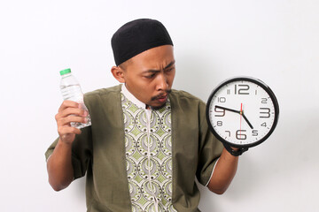 An Indonesian Muslim man in koko and peci holds a mineral water bottle and a clock, looking...