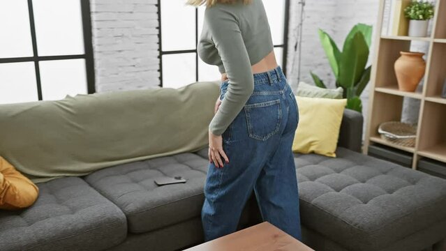 A blonde woman wearing jeans and a green top relaxing on a gray couch inside a cozy living room