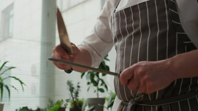 Cropped shot of unrecognizable male chef in striped apron using musat while sharpening knife at kitchen before start cooking