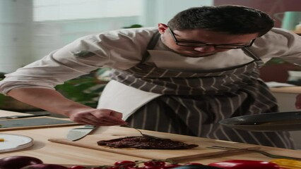 Vertical medium shot of Caucasian chef pouring melted butter from frying pan onto cooked red meat steak lying on wooden cutting board at cooking table - Powered by Adobe