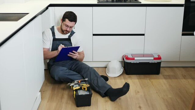 Hardworking man in professional uniform entering data in notepad by hand while sitting on parquet of renovated kitchen. Relaxed labourer analyzing installed plumbing among equipment and headwear.