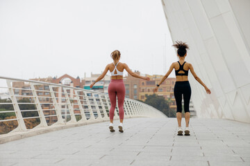 Back view of two sporty women making jumping exercises outside while training together