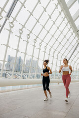 Two active women athlete running side by side along an outdoor track on modern buildings background