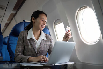 Attractive Asian female passenger on airplane sitting in comfortable seat using laptop and credit...