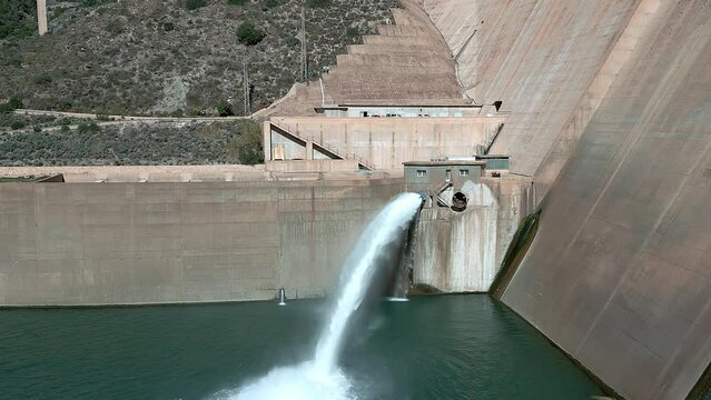 Dam and water reservoir. Howell bunger valve in operation. Dam discharging water. Panoramic aerial view of the dam and hydroelectric plant rising. Dolly out aerial view. Renewable energies.