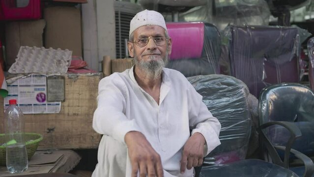 An old man or aged Indian Asian bearded Muslim male in traditional attire wearing eyeglasses sitting outside the furniture store or shop looking at the camera in a busy market or bazaar