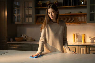 A focused young woman stands in a warmly lt kitchen, gently wiping the countertop with a blue...