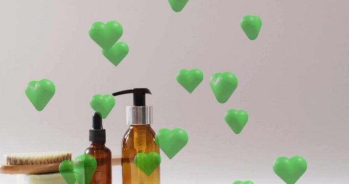 Animation of green hearts rising over soap, brush, self care and beauty products on beige background