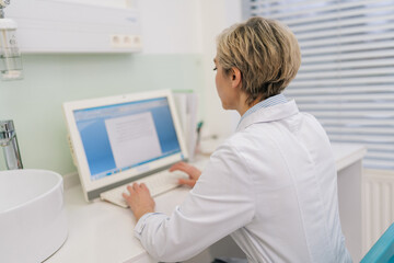Rear view of unrecognizable middle-aged woman general practitioner in white coat using laptop...