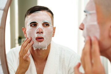 Handsome young man applying sheet mask near mirror in bathroom