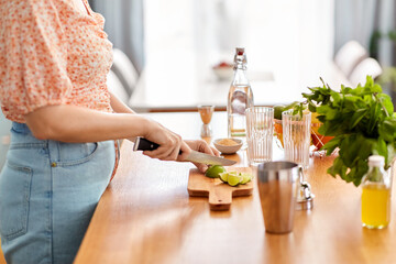 drinks and people concept - close up of woman cutting lime with knife and making cocktail at home kitchen