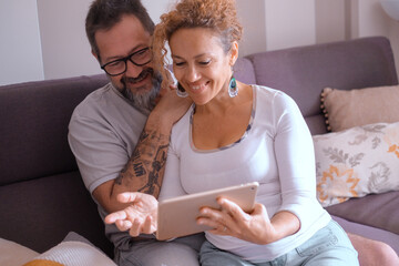 Happy mature Caucasian couple sitting on home sofa using digital tablet browsing the net,...