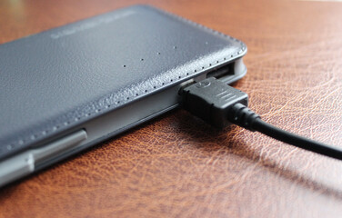 Black USB cable is connected to a discharged power bank with leather upholstery closeup angle view