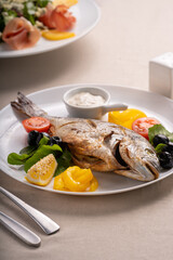 Grilled dorado fish with tomatoes and lemons on a white plate in a restaurant with kitchen utensils