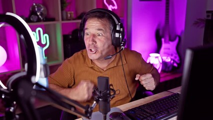 A mature man in headphones expresses excitement at a microphone in a neon-lit gaming room
