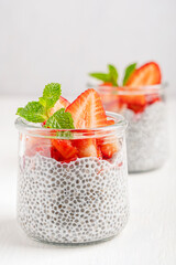 Sweet pudding made from chia seeds with alternative plant based milk topped off with fresh juicy raw strawberries decorated with green mint leaf served in glass jar on white wooden table as dessert