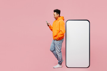 Full body side view fun young man of African American ethnicity he wear yellow hoody casual clothes big huge blank screen mobile cell phone with area use smartphone isolated on plain pink background.