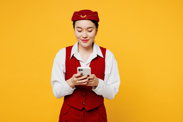 Young smiling happy stewardess flight attendant woman of Asian ethnicity she wear red vest shirt hat use mobile cell phone isolated on plain yellow background studio portrait. Air flight trip concept.