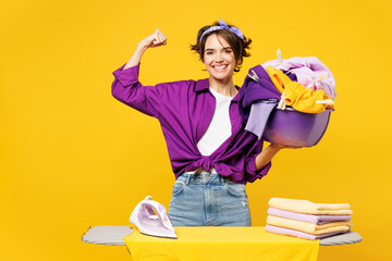 Young woman wear purple casual shirt do housework tidy up ironing clean clothes on board hold basin...