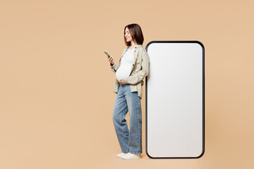 Full body young pregnant woman future mom wear grey shirt with belly stomach tummy baby big blank screen mobile cell phone use smartphone isolated on plain beige background. Maternity family concept.