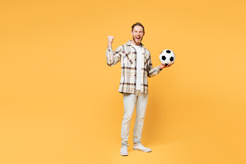 Full body young happy man fan wears brown shirt casual clothes cheer up support football sport team hold in hand soccer ball watch tv live stream do winner gesture isolated on plain yellow background.