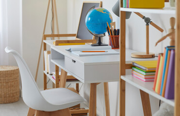 Beautiful modern children's room at home. Kids' study room with white working desk, chair, wooden shelves, books, pencils, notebooks, lamps, Earth globe and chalkboard