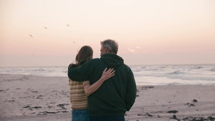 Camera pushes in on rear view of retired senior couple on vacation standing by classic convertible sports car at beach watching morning sunrise on road trip  - shot in slow motion