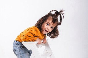 Portrait of a two-year-old girl standing on a ladder with a surprised face on white background