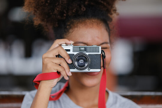Stylish young African American woman taking pictures with a retro camera. Brunette with curly hair in brown leather jacket poses against background of blurred brick building. Close up.