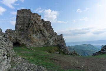 Fototapeta premium The ruins of a medieval fortress on top of a huge rock. Stone walls are a continuation and are combined with rocks. Clear blue sky and clouds. Landscape of nearby mountains and villages. Kojori
